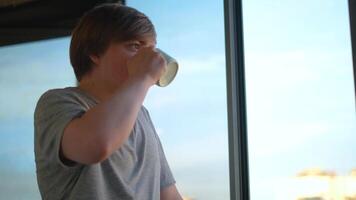 Young man drinks cup of coffee looking out window. Concept. Student drinks coffee in morning looking at morning outside window. Hot coffee in morning with beautiful view from window video