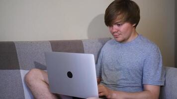 Student looks at laptop while sitting on couch. Concept. Young man smiles while sitting at laptop at home. Social networks and Internet addiction video