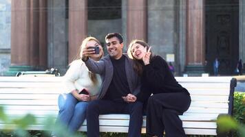 três amigos levando uma selfie juntos enquanto sentado em uma Banco às parque. meios de comunicação. dois meninas e uma homem sorridente alegremente e desfrutando Tempo junto. video