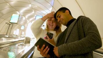 copains bavardage sur le escaliers mécaniques de Publique souterrain transport. médias. bas vue de homme et femme avec une téléphone intelligent et une tablette discuter quelque chose. video