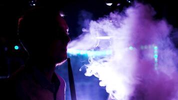 Side view of male silhouette in neon light smoking hookah, relaxing at night club. Media. Man smoking shisha, exhaling thick smoke from his mouth. video