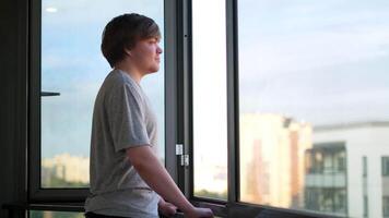 An adult man in a grey t shirt standing against an opened window at the balcony and enjoying the view. Concept. Side view of a happy man looking into the new day through the opened window at home. video