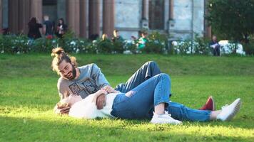 Beautiful young couple lie and laugh on grass in park. Concept. Couple in love embrace lying on green grass in sun. Young man and woman laughing and hugging on grass in park video