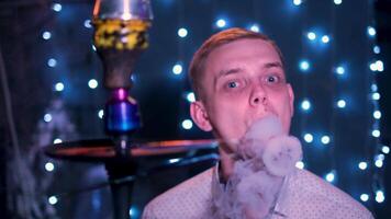 Young blond man with blue eyes blowing smoke rings. Media. Male smoking hookah at the night bar, exhaling smoke on blue shining lights background. video