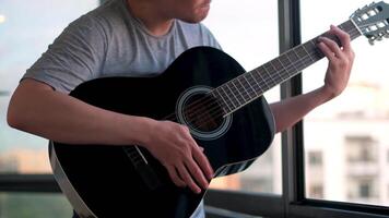 Man standing on the balcony against windows and playing guitar. Concept. Young guy spending time at home practicing in playing acoustic guitar, social distancing and covid 19 concept. video