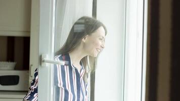 Dark haired smiling woman opens the door of a balcony and looks outside. Video. Side view of a happy lady at home looking outdoors through a window of a living room on a sunny day. video