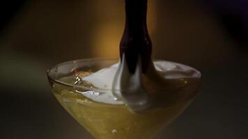 Putting cream to alcohol yellow cocktail with ice. Stock footage. Close up of adding to a cocktail in the martini glass a whipped cream on on dark blurred background. video