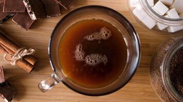 Morning hot beverage preparation. Concept. Top view of woman hands putting a spoon of instant coffee and a sugar cube into the transparent cup on a wooden table background with chocolate and cinnamon. video