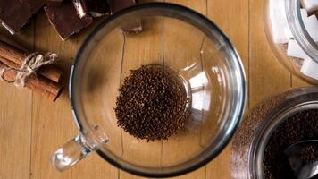 Morning hot beverage preparation. Concept. Top view of woman hands putting a spoon of instant coffee and a sugar cube into the transparent cup on a wooden table background with chocolate and cinnamon. video