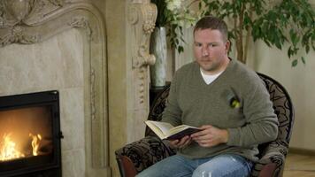 Man sitting in a chair,reading a book in front of fireplace. Video. Butterfy flying near the male wearing pullover and jeans sitting at home by the fire and reading. video