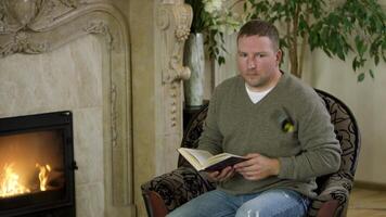 Man sitting in a chair,reading a book in front of fireplace. Video. Butterfy flying near the male wearing pullover and jeans sitting at home by the fire and reading. video