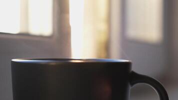 ein Tasse von heiß Kaffee oder Tee und das Dampf steigend oben im das Luft. Konzept. schließen oben von schwarz Tasse mit heiß Getränk Stehen im Vorderseite von das Fenster beim Zuhause auf verschwommen Hintergrund. video