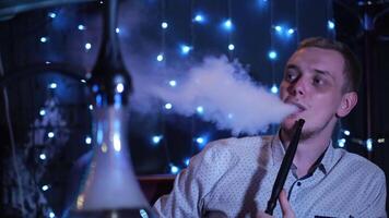 Portrait of a stylish blond adult guy smoking hookah indoors in a cafe. Media. Male with big blue eyes sitting at the table and smoking shisha, concept of leisure. video