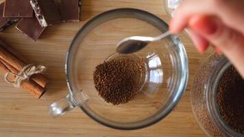 Close up top view of a woman hand putting granulated instant coffee, sugar, and hot water into the mug. Concept. Making transparent cup of coffee placed on brown wooden surface with chocolate and video