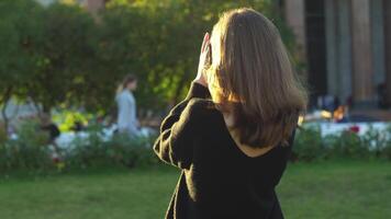 Rückseite Aussicht von Frau im schwarz Sweatshirt halten Fotoapparat und nehmen Foto beim Sommer- Grün Park. Medien. jung Mädchen Fotografieren Sehenswürdigkeiten im ein groß Stadt im das Sommer. video