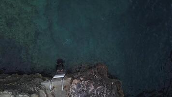 hombre consiguiendo fuera de profundo azul mar. existencias. parte superior aéreo ver de un masculino nadador en negro nadando traje alpinismo escalera fuera de agua en pedregoso costa. video