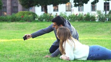 glücklich Paar nehmen Selfie auf Smartphone und lächelnd während Lügen auf Gras im Park. Medien. Seite Aussicht von jung Mann und Frau haben Spaß im das Sommer. video