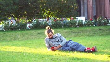 A young man is browsing on his smartphone while lying on nature on green grass in the summertime. Concept. Serious man using mobile phone while relaxing in a city park, lying on the grass, chatting video