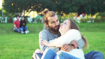 retrato de contento encantador caucásico romántico Pareja sentado en césped y abrazando cada otro en parque. concepto. joven hombre y mujer sentado en el césped y disfrutar en el abierto aire, masculino besos un niña en video