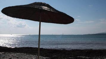 A straw umbrella on a beautiful pebble beach. Concept. Summer straw umbrella and the reflection of the sun in rippled water surface, summer holiday concept. video