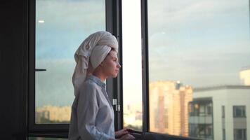 Young woman in a shirt meets the morning on the balcony, daily routine. Concept. Yong girl with a towel on her head standing on the balcony in the morning and looking sad and tired. video