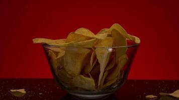 Close up of potato chips inside the transparent glass bowl. Stock footage. Crispy junk product in a small deep dish lighted isolated on red background. video