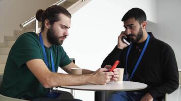 deux les hommes d'affaires séance à le table pendant café Pause et en utilisant leur téléphones intelligents. médias. Jeune homme à la recherche concentré tandis que navigation navigation sur le l'Internet et un autre gars parlant sur mobile video