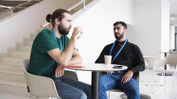 Two serious young people drinking coffee and talking inside the building with white walls and stairs. Media. Confident business partners discussing ideas, planning projects and startups. video