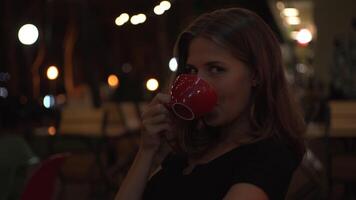 Beautiful young female at cafe drinking coffee or tea, looking into the camera and smiling. Media. Portrait of a flirting woman drinking hot beverage at night summer cafe. video