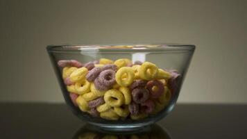 Concept of food and breakfast. Stock footage. Close up of colorful ring corn flakes in a transparent glass bowl on dark table surface and grey wall background. video