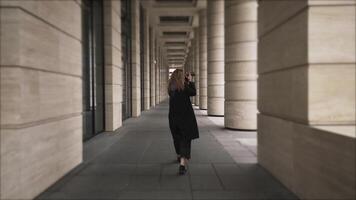 une fille en marchant parmi Colonnes avec sa retour tourné à caméra. action. arrière vue de une femme avec frisé cheveux en marchant en plein air près le bâtiment avec gros les fenêtres et géant piliers. video