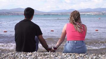 Rear view of man and woman holding hands with two glasses of red wine standing near on the pebble beach. Media. Concept of honeymoon, woman with braids and a man enjoying summer marine landscape. video