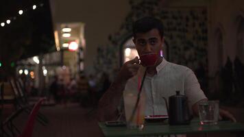 Young pensive man sitting alone with a cup of hot coffee or tea at the outdoor terrace late in the evening. Media. Sad male in white shirt waiting for someone alone at the street cafe. video