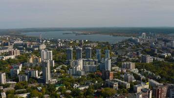 City with high-rise buildings on background of lake and green forest. Stock footage. Modern buildings in beautiful green city with lake and forest horizon. Top view of panorama of modern city in video