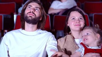 familia acecho película en cine. medios de comunicación. retrato de contento mamá, papá, y cura pequeño niña sentado juntos y acecho un interesante película, concepto de ocio. video