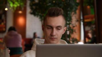 Young man is working on laptop in cafe and talking on phone. Stock footage. Young freelancer is working on laptop and answering calls. Remote work with laptop and cafe calls video