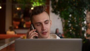 Young man is working on laptop in cafe and talking on phone. Stock footage. Young freelancer is working on laptop and answering calls. Remote work with laptop and cafe calls video