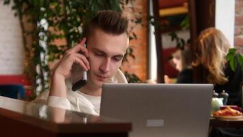 joven hombre es hablando en teléfono a ordenador portátil en cafetería. valores imágenes. joven hombre es hablando en teléfono mientras trabajando en ordenador portátil. hombre respuestas trabajo llamadas mientras trabajando en ordenador portátil en café video