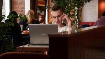 joven hombre es hablando en teléfono a ordenador portátil en cafetería. valores imágenes. joven hombre es hablando en teléfono mientras trabajando en ordenador portátil. hombre respuestas trabajo llamadas mientras trabajando en ordenador portátil en café video