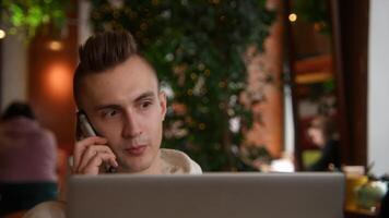 giovane uomo è Lavorando su il computer portatile nel bar e parlando su Telefono. azione filmato. giovane libero professionista è Lavorando su il computer portatile e segreteria chiamate. a distanza opera con il computer portatile e bar chiamate video