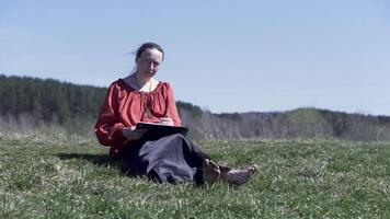 Frau Skizzierung im ein Park Sitzung auf das kurz Grün Gras unter das Sommer- Sonnenlicht. Clip. weiblich im traditionell Russisch Hemd und lange Rock Zeichnung draußen, inspirierend Natur. video