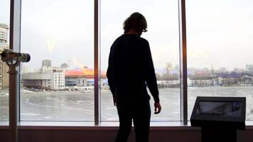 un hombre mira a un hermosa ver de el ciudad y un congelado lago desde el observación cubierta. medios de comunicación. posterior ver de un joven hombre dentro el edificio con vaso paredes disfrutando invierno paisaje. video