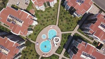 Top aerial view of a swimming pool located outdoors near several residential houses. Clip. Red roofs of houses contrasting to green lawn and blue water of a pool. video