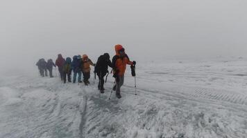 Mountaineering activity on the steep mountain slope. Clip. Group of extreme tourists walking one by one on icy surface of a hill in foggy cold weather. video