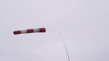 windsock geblazen door de wind Aan bewolkt, grijs, zwaar lucht achtergrond. actie. verlaten meteorologie windsock van rood en wit kleur zwaaiend in de sterk wind. video