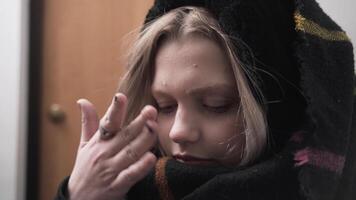 Portrait of a young blond girl standing dressed in a hallway at home. Action. Woman with a warm scarf on her head looking down, laughing, and touching her nose in front of a wooden door. video