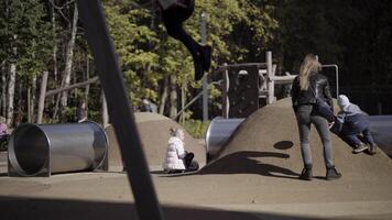 al aire libre moderno niños patio de recreo con diferente equipo para divertido y para Deportes actividades. acción. un niña es balanceo y un madre con su hijo son molesto a conquistar el artificial colina. video