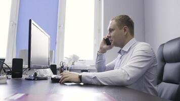 Businessman working in his office in front of computer screen. Action. Side view of male in a shirt talking on his mobile phone about work. video