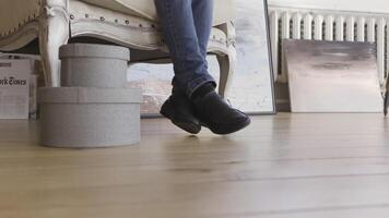 Legs of a child in black shoes and blue jeans indoors in a room. Action. Child sitting in a white armchair with a round shaped present boxes standing near. video