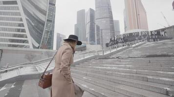 Beautiful business lady in a brown coat and black hat going upstairs in the modern city district with skyscrapers. Action. Side view of a woman walking near beautiful glass facade buildings. video